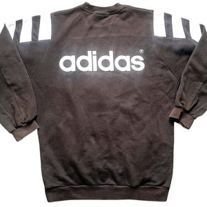 May include: A brown sweatshirt with white stripes on the shoulders and the word "adidas" in white letters on the front.
