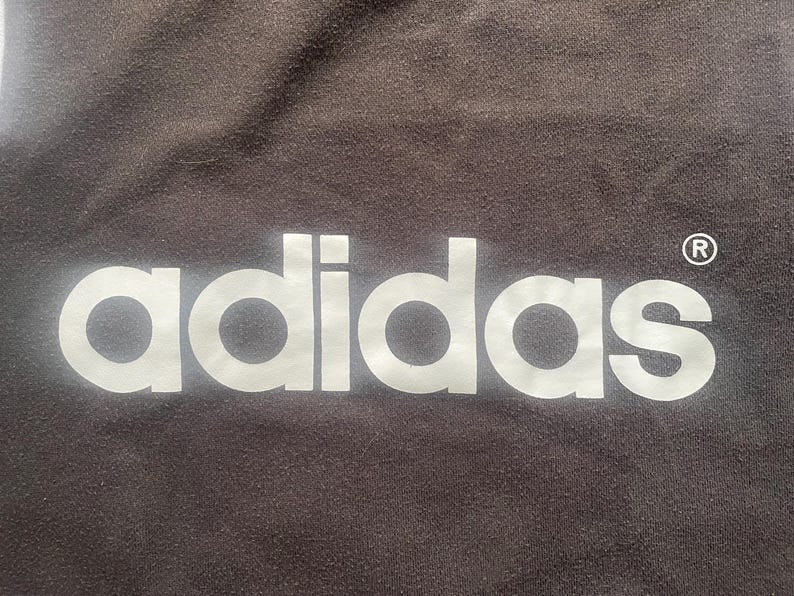 May include: White adidas logo on a dark brown background with a registered trademark symbol.