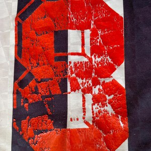 Pode incluir: Um close-up de um n&uacute;mero 8 vermelho impresso em um tecido listrado branco e preto. O n&uacute;mero est&aacute; ligeiramente desbotado e rachado, dando-lhe um visual vintage.
