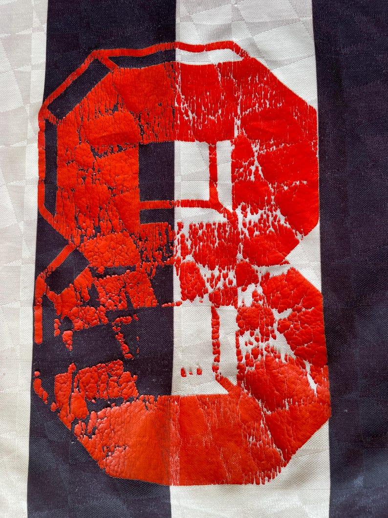 Pode incluir: Um close-up de um n&uacute;mero 8 vermelho impresso em um tecido listrado branco e preto. O n&uacute;mero est&aacute; ligeiramente desbotado e rachado, dando-lhe um visual vintage.