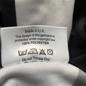 K&ouml;nnte beinhalten: Nahaufnahme eines wei&szlig;en Etiketts mit schwarzem Text, eingen&auml;ht in einen schwarz-wei&szlig; gestreiften Stoff. Das Etikett lautet "Made in U.K." und enth&auml;lt Waschanweisungen. Das Kleidungsst&uuml;ck besteht aus 100 % Polyester.