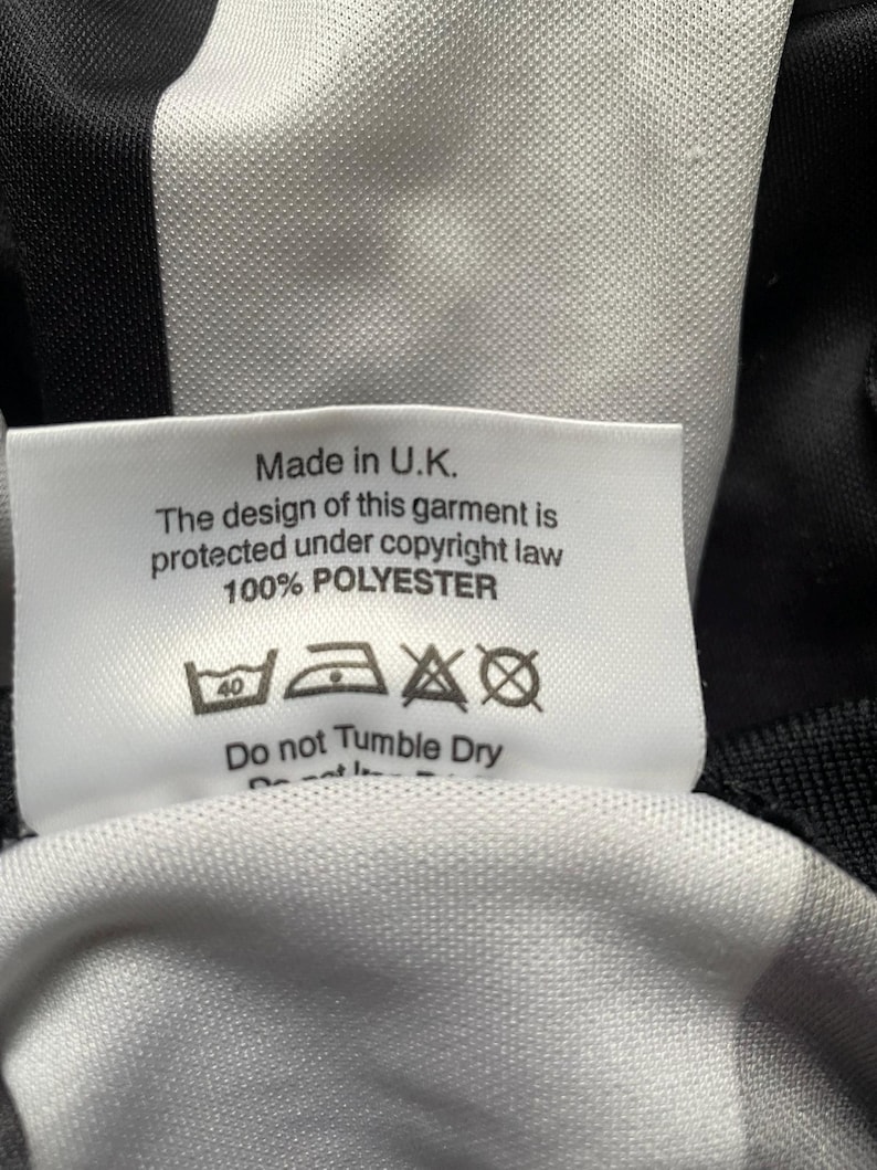 K&ouml;nnte beinhalten: Nahaufnahme eines wei&szlig;en Etiketts mit schwarzem Text, eingen&auml;ht in einen schwarz-wei&szlig; gestreiften Stoff. Das Etikett lautet "Made in U.K." und enth&auml;lt Waschanweisungen. Das Kleidungsst&uuml;ck besteht aus 100 % Polyester.