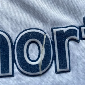 May include: Close-up of a white fabric with the word "nort" in navy blue lettering with a white outline. The "o" in the word is slightly torn.