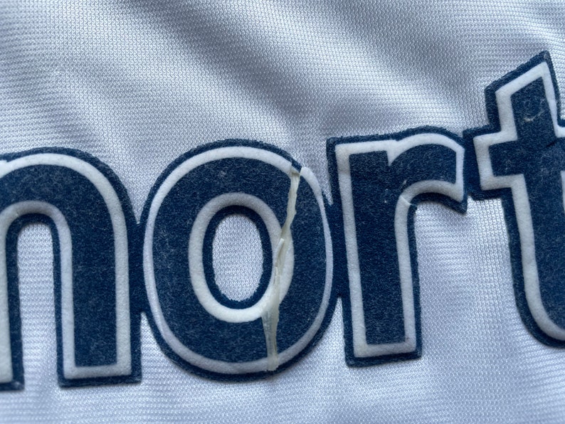 May include: Close-up of a white fabric with the word "nort" in navy blue lettering with a white outline. The "o" in the word is slightly torn.
