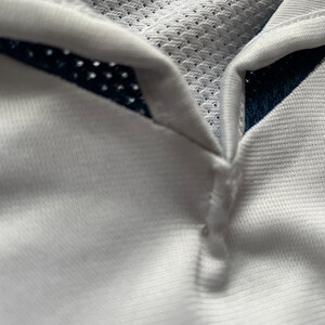 May include: White athletic shirt with a navy blue mesh panel on the collar. The shirt has a button closure at the neck.