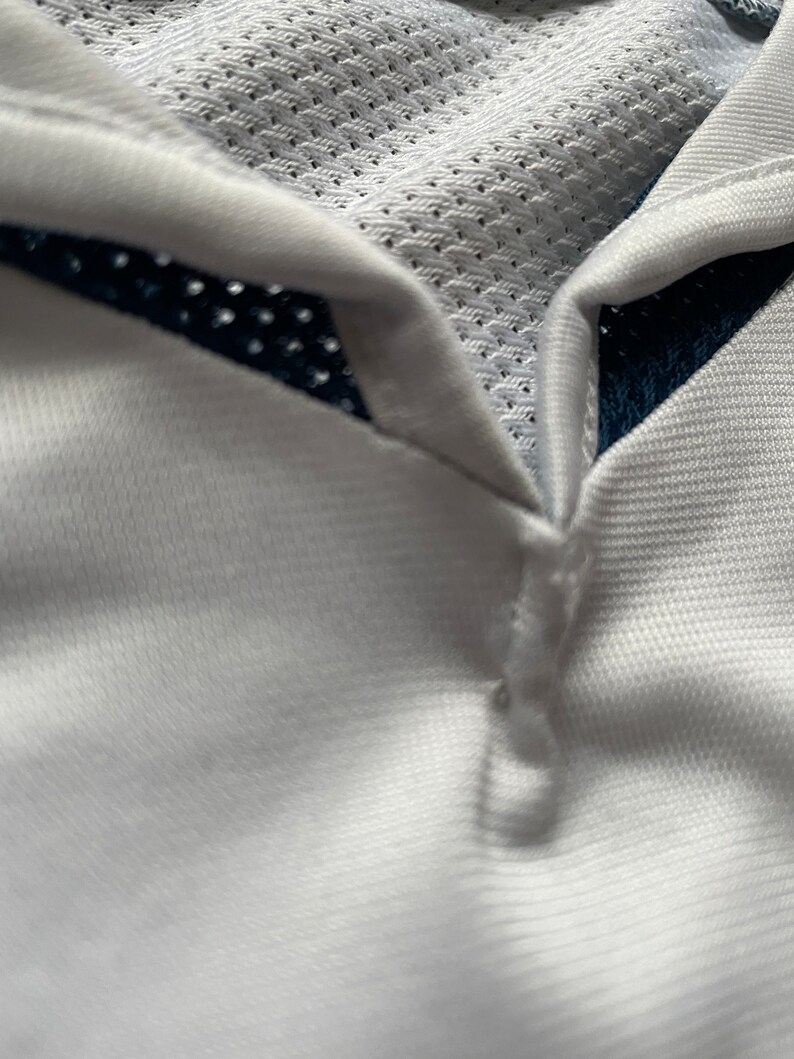 May include: White athletic shirt with a navy blue mesh panel on the collar. The shirt has a button closure at the neck.