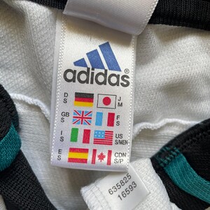 May include: A close-up of a white Adidas label featuring the brand's blue logo. The label displays size and country codes, plus the numbers 635825 and 16593. The garment has black and teal detailing.