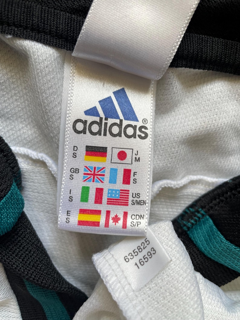 May include: A close-up of a white Adidas label featuring the brand's blue logo. The label displays size and country codes, plus the numbers 635825 and 16593. The garment has black and teal detailing.
