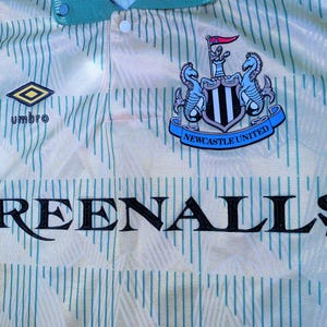 K&ouml;nnte beinhalten: Ein wei&szlig;es und gr&uuml;nes Fu&szlig;balltrikot mit dem Wappen von Newcastle United und dem Wort "GREENALLS" auf der Vorderseite. Das Trikot hat einen gr&uuml;nen Kragen und ein rautenf&ouml;rmiges Logo mit dem Wort "umbro" darunter.
