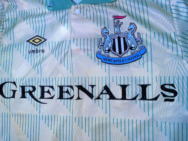 K&ouml;nnte beinhalten: Ein wei&szlig;es und gr&uuml;nes Fu&szlig;balltrikot mit dem Wappen von Newcastle United und dem Wort "GREENALLS" auf der Vorderseite. Das Trikot hat einen gr&uuml;nen Kragen und ein rautenf&ouml;rmiges Logo mit dem Wort "umbro" darunter.