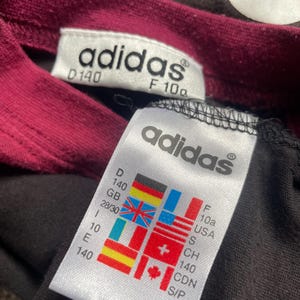 Puede incluir: Primer plano de una camiseta Adidas negra con cuello burdeos. La camiseta tiene dos etiquetas blancas con el logo de Adidas e informaci&oacute;n de tallas, incluidas medidas en cm y conversiones de tallas internacionales.