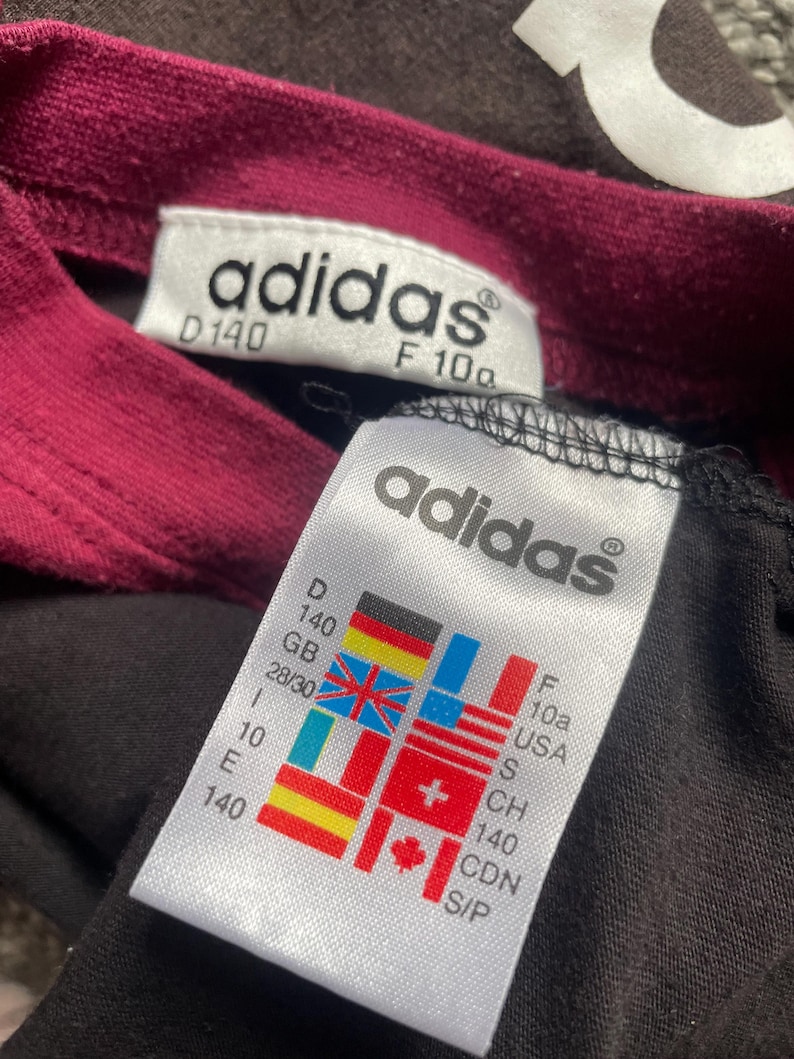 Puede incluir: Primer plano de una camiseta Adidas negra con cuello burdeos. La camiseta tiene dos etiquetas blancas con el logo de Adidas e informaci&oacute;n de tallas, incluidas medidas en cm y conversiones de tallas internacionales.
