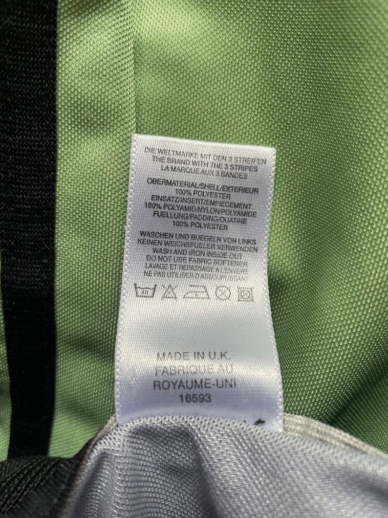 May include: Close-up of a tag with text in multiple languages, including English, French, and German. The tag is white with black text and is attached to a green and black fabric. The tag indicates the item was made in the U.K.
