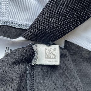 Pode incluir: Close-up de uma etiqueta de roupa preta e branca com um c&oacute;digo de barras e o n&uacute;mero 7115500101066.