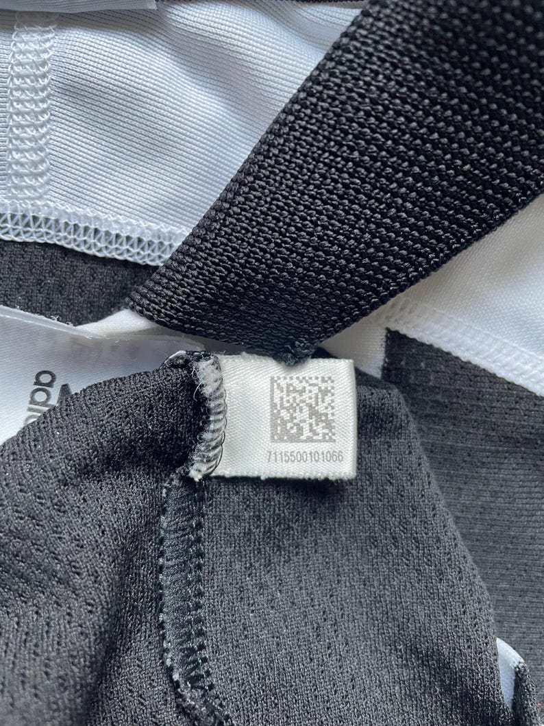 Pode incluir: Close-up de uma etiqueta de roupa preta e branca com um c&oacute;digo de barras e o n&uacute;mero 7115500101066.