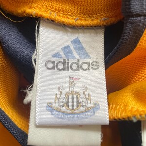 May include: White tag with black text that reads "adidas" and a blue and white crest with the text "Newcastle United".