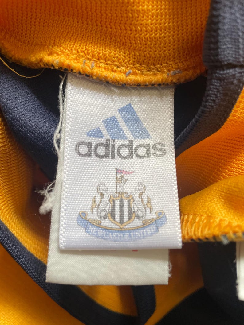 May include: White tag with black text that reads "adidas" and a blue and white crest with the text "Newcastle United".
