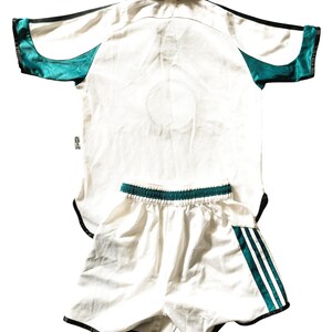 Pu&ograve; includere: Maglia da calcio bianca con bordini neri e verdi e pantaloncini bianchi abbinati con strisce verdi e nere. La maglia ha un colletto nero.