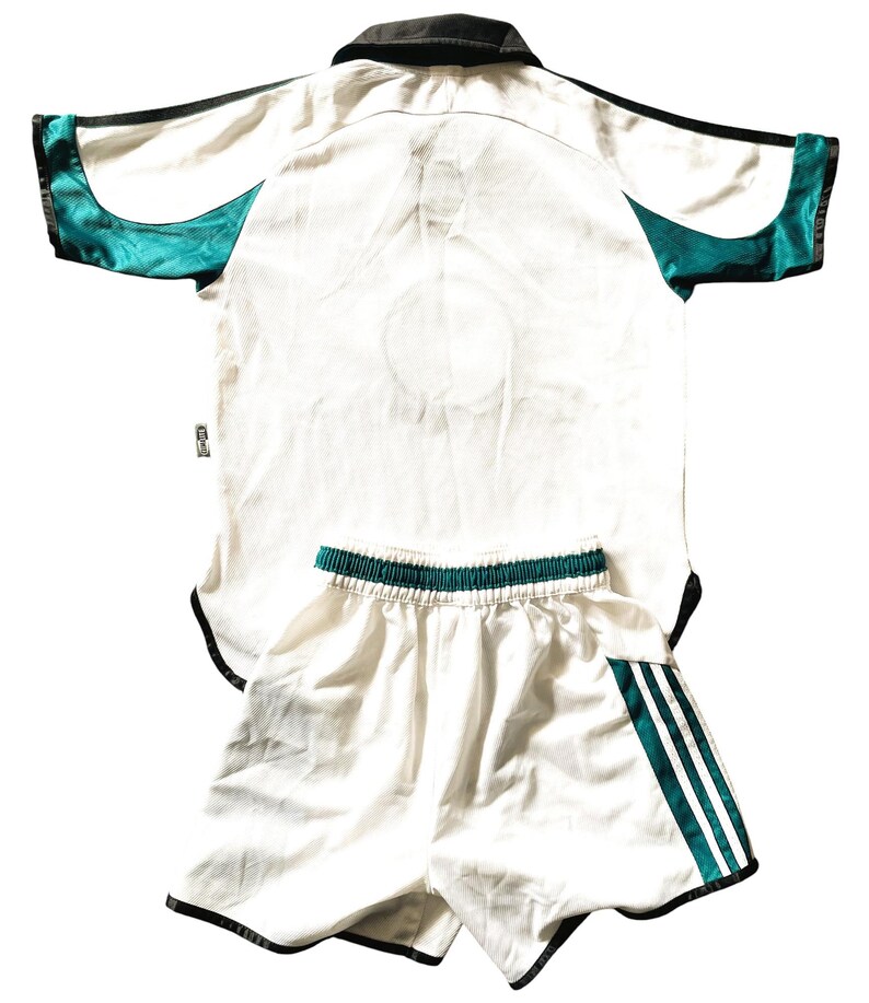 Pu&ograve; includere: Maglia da calcio bianca con bordini neri e verdi e pantaloncini bianchi abbinati con strisce verdi e nere. La maglia ha un colletto nero.