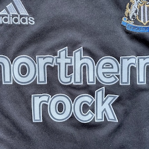 May include: Black soccer jersey with the Adidas logo, a team crest, and the words "northern rock" in a silver-grey color.