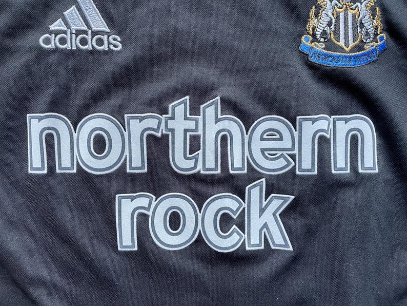 May include: Black soccer jersey with the Adidas logo, a team crest, and the words "northern rock" in a silver-grey color.