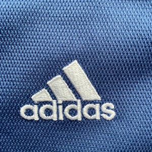Op de afbeelding: Een close-up van een blauwe stof met een wit geborduurd Adidas-logo.