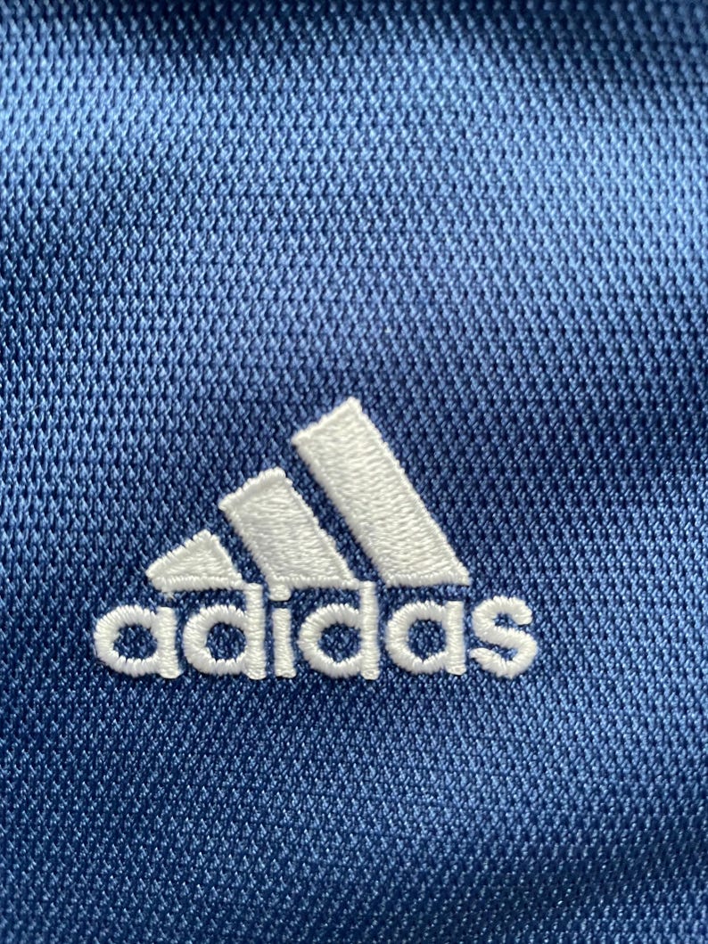 Op de afbeelding: Een close-up van een blauwe stof met een wit geborduurd Adidas-logo.