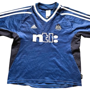 Op de afbeelding: Een blauw en zwart voetbalshirt met witte strepen op de schouders. Het shirt heeft een V-hals en de tekst "ntl:" in wit. Het Adidas-logo is zichtbaar, samen met een teamembleem.