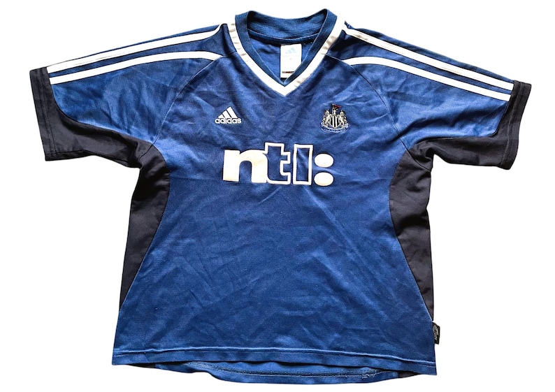 Op de afbeelding: Een blauw en zwart voetbalshirt met witte strepen op de schouders. Het shirt heeft een V-hals en de tekst "ntl:" in wit. Het Adidas-logo is zichtbaar, samen met een teamembleem.