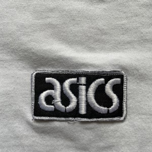 May include: White t-shirt with a black and white embroidered patch that says "asics" on it.