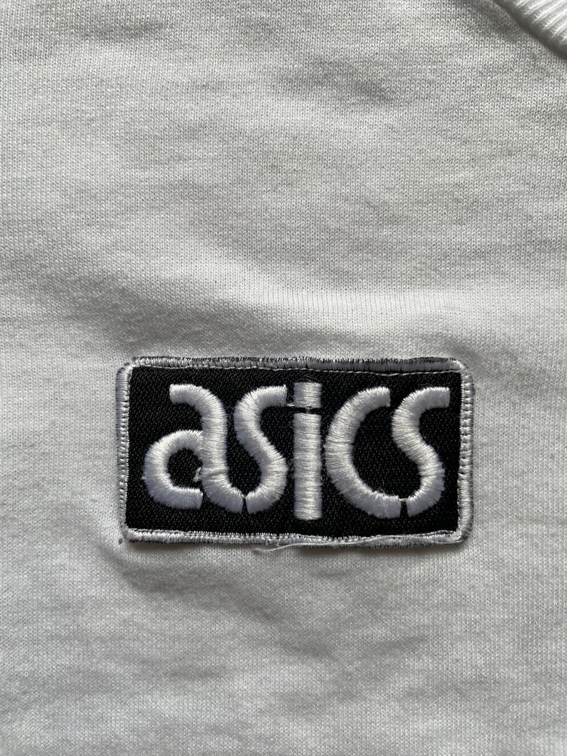 May include: White t-shirt with a black and white embroidered patch that says "asics" on it.