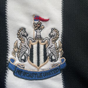 May include: Close-up of a black and white striped fabric featuring an embroidered crest. The crest includes a shield with a flag, two seahorses, and the words "Newcastle United" on a blue banner.