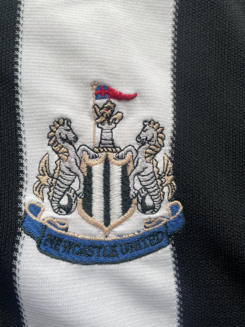 May include: Close-up of a black and white striped fabric featuring an embroidered crest. The crest includes a shield with a flag, two seahorses, and the words "Newcastle United" on a blue banner.