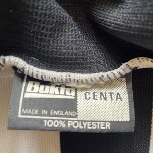 Może przedstawiać: Zbliżenie czarno-białego pasiaka z metką. Na metce widnieje napis "Bukta" i "CENTA", z nadrukiem "MADE IN ENGLAND" i "100% POLYESTER" poniżej. Tkanina wydaje się być materiałem dżersejowym.