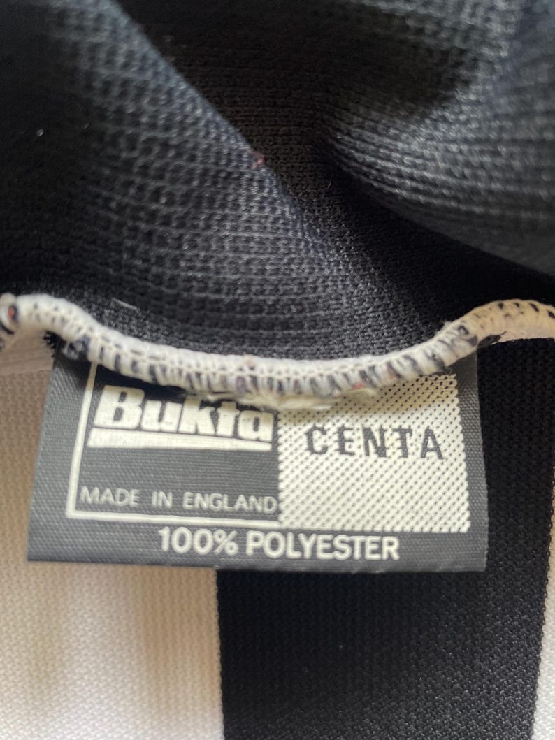 Może przedstawiać: Zbliżenie czarno-białego pasiaka z metką. Na metce widnieje napis "Bukta" i "CENTA", z nadrukiem "MADE IN ENGLAND" i "100% POLYESTER" poniżej. Tkanina wydaje się być materiałem dżersejowym.