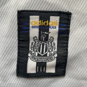 Puede incluir: Un parche en blanco y negro con el logotipo de Adidas y las palabras "OFFICIAL REPLICA" en dorado. El parche tambi&eacute;n presenta el escudo y el texto del Newcastle United.