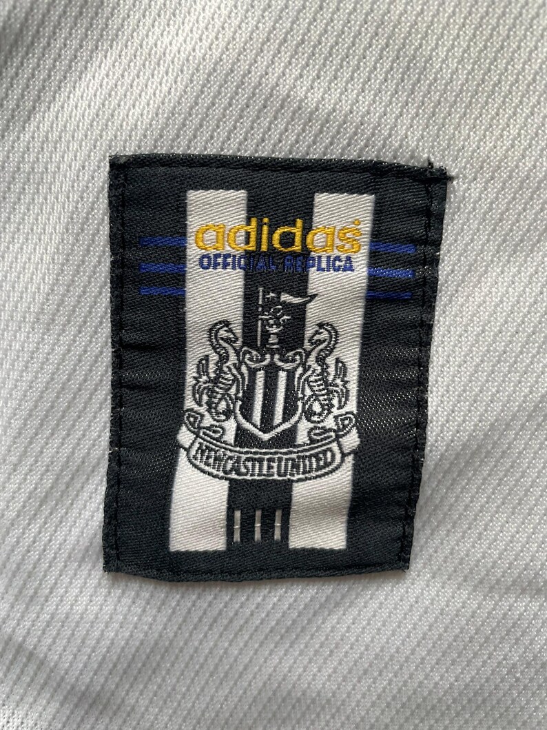 Puede incluir: Un parche en blanco y negro con el logotipo de Adidas y las palabras "OFFICIAL REPLICA" en dorado. El parche tambi&eacute;n presenta el escudo y el texto del Newcastle United.