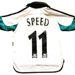 Puede incluir: Camiseta de f&uacute;tbol blanca con detalles en negro, que muestra el nombre "SPEED" y el n&uacute;mero "11" en negro. La camiseta tiene detalles de malla verde azulado en las mangas y un cuello negro. El logotipo de la Premier League es visible.