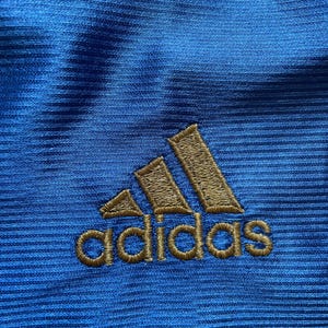 K&ouml;nnte beinhalten: Nahaufnahme eines blauen Stoffes mit einem goldenen gestickten Adidas-Logo.