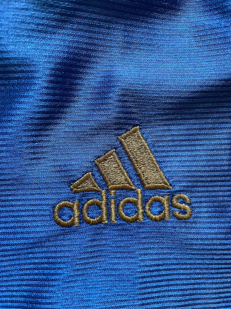 K&ouml;nnte beinhalten: Nahaufnahme eines blauen Stoffes mit einem goldenen gestickten Adidas-Logo.