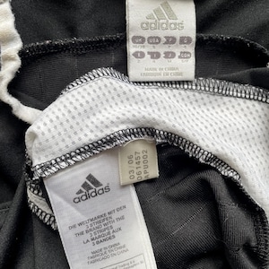 Pu&ograve; includere: Primo piano di abbigliamento sportivo Adidas nero con dettagli bianchi. L'immagine mostra pi&ugrave; etichette con il logo Adidas e testo in inglese, francese e spagnolo. Le etichette includono informazioni sulle taglie e sulla produzione.