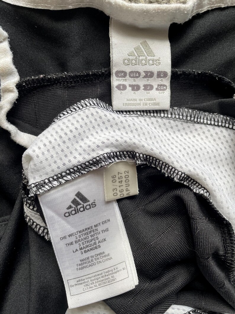 Pu&ograve; includere: Primo piano di abbigliamento sportivo Adidas nero con dettagli bianchi. L'immagine mostra pi&ugrave; etichette con il logo Adidas e testo in inglese, francese e spagnolo. Le etichette includono informazioni sulle taglie e sulla produzione.
