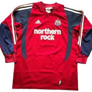 Pu&ograve; includere: Una maglia da calcio a maniche lunghe rossa con dettagli bianchi e blu. La maglia ha il logo Adidas, lo stemma del Newcastle United e il testo "northern rock" sul davanti.