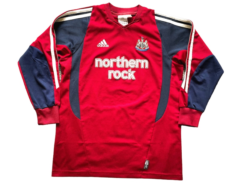Pu&ograve; includere: Una maglia da calcio a maniche lunghe rossa con dettagli bianchi e blu. La maglia ha il logo Adidas, lo stemma del Newcastle United e il testo "northern rock" sul davanti.