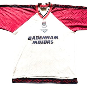 May include: A vintage white soccer jersey with red sleeves and black trim. The jersey features the text "DAGENHAM MOTORS" in blue, along with a team logo and the "PONY" logo. The sleeves have a geometric pattern.