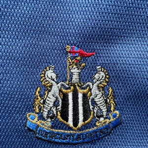 Op de afbeelding: Een blauwe geborduurde patch met een zwart-wit schild, twee gouden zeepaardjes en een gouden banner met de tekst "NEWCASTLE UNITED".