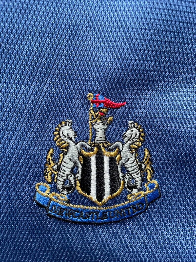 Op de afbeelding: Een blauwe geborduurde patch met een zwart-wit schild, twee gouden zeepaardjes en een gouden banner met de tekst "NEWCASTLE UNITED".
