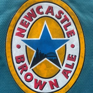 May include: A close-up of a circular logo with the words "Newcastle Brown Ale" in a white circle with a red outline. The circle is surrounded by a yellow circle with a red outline. A blue star with a cityscape inside is in the center of the logo.