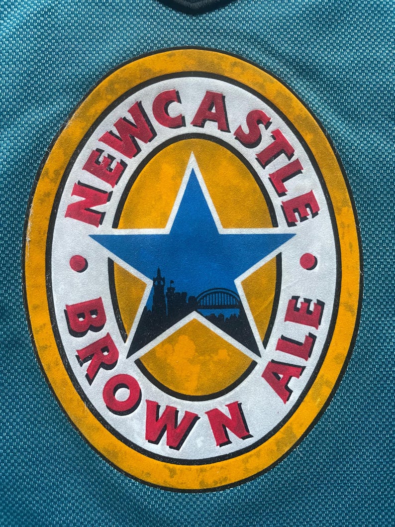 May include: A close-up of a circular logo with the words "Newcastle Brown Ale" in a white circle with a red outline. The circle is surrounded by a yellow circle with a red outline. A blue star with a cityscape inside is in the center of the logo.