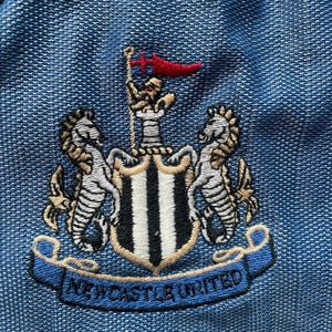 May include: Embroidered Newcastle United crest on a blue fabric. The crest features a shield with black and white stripes, flanked by two seahorses, and a banner that reads "NEWCASTLE UNITED". A red flag is at the top.